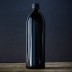 Botella alta de vidrio Miron Violeta sobre superficie de madera con fondo oscuro.