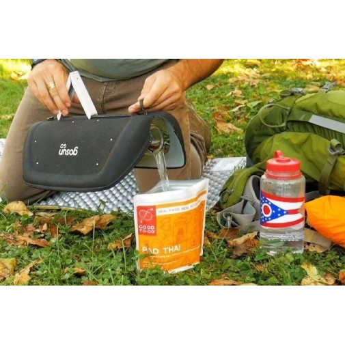 Persona vertiendo agua en una bolsa de comida Pad Thai al aire libre, rodeada de equipos de cocina solar.