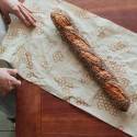 Persona envolviendo una baguette fresca con semillas en papel estampado ecológico sobre una mesa de madera.