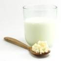 Un vaso de leche al lado de una cuchara de madera con gránulos de Kefirko sobre un fondo blanco.