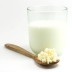 Un vaso de leche al lado de una cuchara de madera con gránulos de Kefirko sobre un fondo blanco.