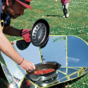 Hombre cocinando en una cocina solar al aire libre mientras los niños juegan en el fondo.
