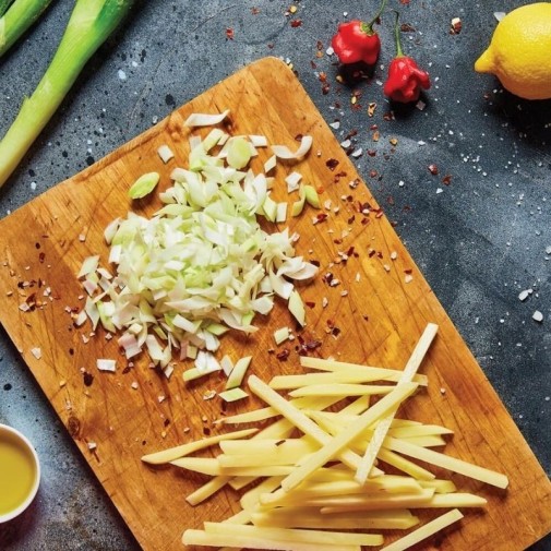 Cebollas y patatas picadas sobre una tabla de madera con frutas y verduras, limón y aceite cerca.