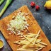 Cebollas y patatas picadas sobre una tabla de madera con frutas y verduras, limón y aceite cerca.