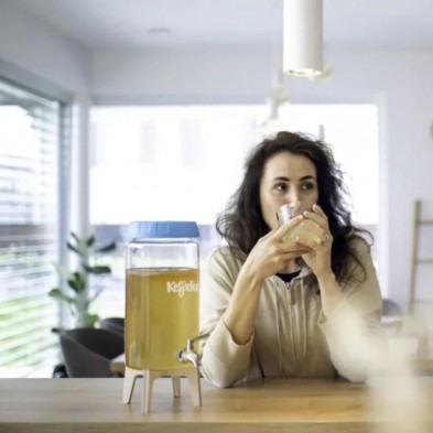 Mujer bebiendo una bebida de un vaso en una mesa con un dispensador de bebidas cerca.