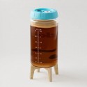 Frasco transparente fermentador de kombucha con tapa azul y marcas de llenado sobre soporte de madera lleno de líquido marrón.