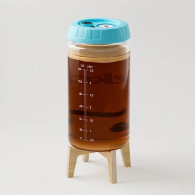 Frasco transparente fermentador de kombucha con tapa azul y marcas de llenado sobre soporte de madera lleno de líquido marrón.