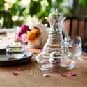 Jarra de vidrio artesanal GALILEO y copas sobre una mesa con pétalos y flores al fondo