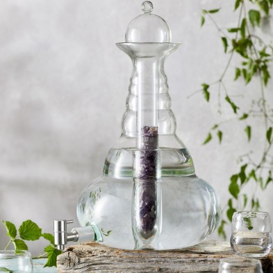 Elegante dispensador de agua de vidrio Beauty de 5L, con cristal en su interior, sobre madera rústica, rodeado de hojas.