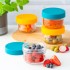 Pequeños frascos de vidrio con tapas de colores, llenos de frutas y verduras frescas en una encimera de cocina.