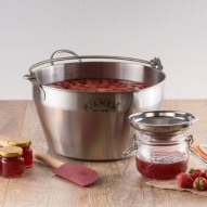 Olla para hacer mermeladas Kilner con fresas, un frasco, un colador, una espátula y frascos de mermelada sobre una mesa.