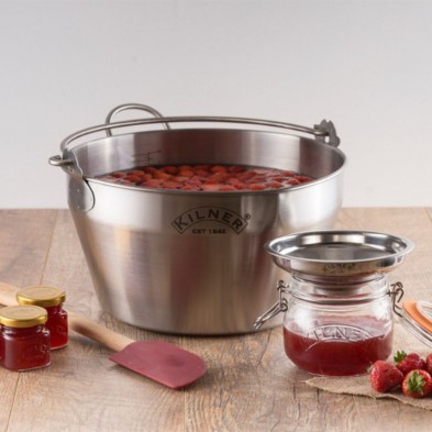Olla para hacer mermeladas Kilner con fresas, un frasco, un colador, una espátula y frascos de mermelada sobre una mesa.