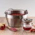 Olla para hacer mermeladas Kilner con fresas, un frasco, un colador, una espátula y frascos de mermelada sobre una mesa.