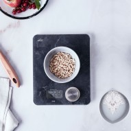 Bol blanco con piñones en una balanza de cocina digital, rodeado de fruta y sal.