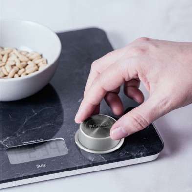 Una mano pulsando el botón econmove de la balanza digital de cocina con un bol de piñones.