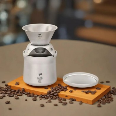 Cafetera de titanio con tapa, sobre una tabla de madera, rodeada de granos de café.
