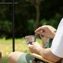Persona sentada al aire libre, removiendo una bebida en la taza de titanio sobre un platillo con cucharilla