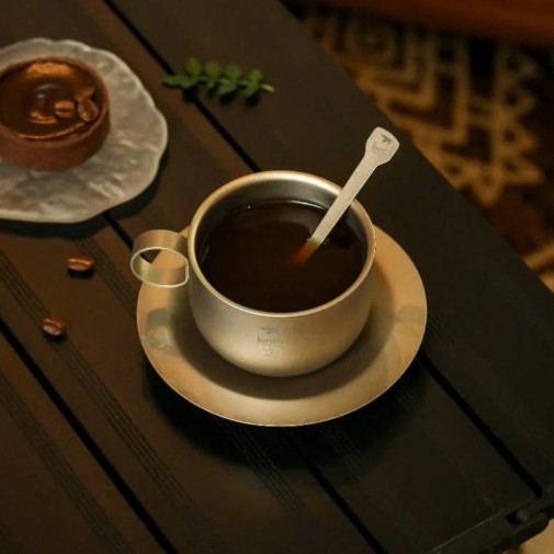 taza de café  de titanio con una cuchara sobre un platillo, junto a un postre en una mesa oscura.