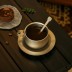 taza de café  de titanio con una cuchara sobre un platillo, junto a un postre en una mesa oscura.
